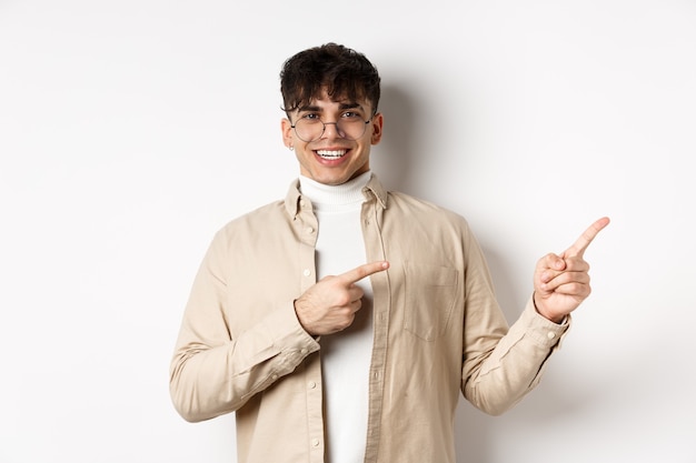 Proud and happy young man in glasses showing logo, pointing fingers at upper right corner and smiling, standing on white wall.