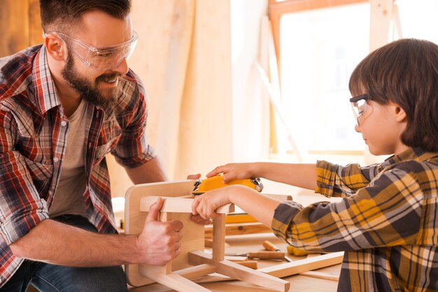 Proud to have a son. Cheerful young male carpenter helping his son tosand wooden chair in workshop