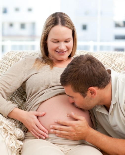 Proud man kissing the belly of his pregnant girlfriend sitting in the sofa