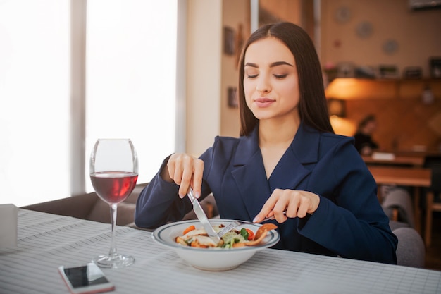 Orgogliosa e simpatica giovane donna sedersi a tavola e mangiare insalata con forchetta e coltello.