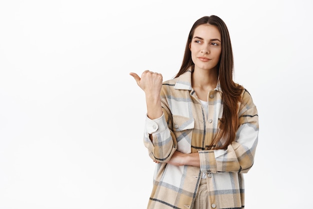 Proud and pleased young woman nod approval and points aside at cool good thing praise nice product or recommend company look left standing over white background