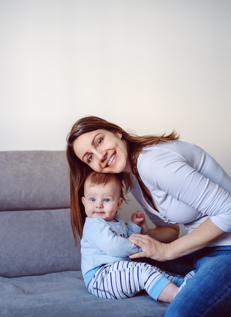 Proud smiling caucasian happy mother posing with her 6 months old adorable baby boy while sitting on sofa in living room.