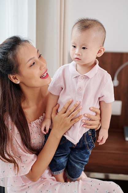 Proud young mother looking at little son standing on her laps and telling him something