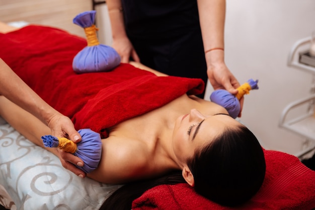 Providing relaxing session. Peaceful young woman lying on massage bed while masters giving her procedure with blue herbal bags