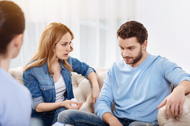 During psychological session. Pleasant cheerless attractive woman looking at her husband and saying something to him while holding a tissue