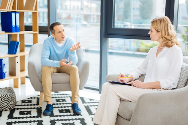 Psychological treatment. Nice professional female psychologist sitting in the armchair and taking notes while listening to her patient