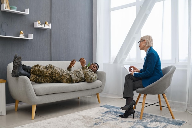 Psychologist making notes on therapy session with sad soldier. War veteran talking lying on couch