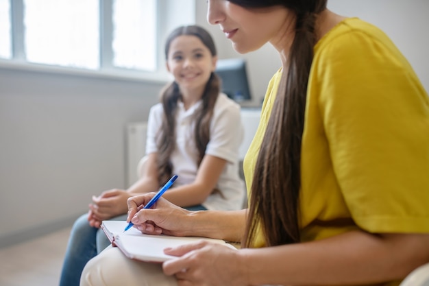 Lavoro psicologo. donna in camicetta gialla che scrive con la penna in taccuino e ragazza allegra che si siede nella stanza luminosa durante il giorno