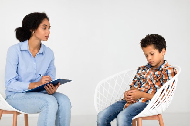 Psychotherapist woman noting as distressed boy sits in thoughts indoors