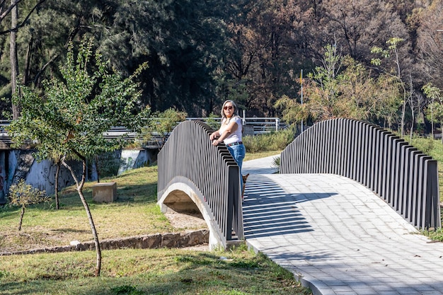 Public park with a woman with her dog standing on bridge over a stream trees with green foliage in background sunglasses casual clothes sunny day in Zapopan Jalisco Mexico