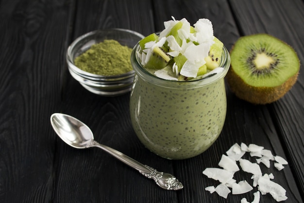 Pudding with chia, matcha tea, kiwi and coconut flakes in the glass jar on the black wooden