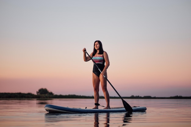 Puddle boarding female person of middle age rowing with oar looking at sun setting on lake with amazing pink sky in background in swimsuit in evening time Active lifestyle for older people