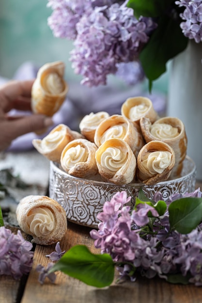 puff pastry horns with vanilla cream in a metal box in spring still life with a bouquet of lilacs on a wooden table