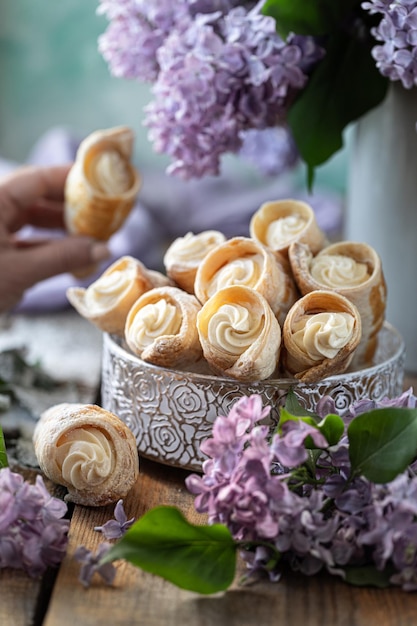 Puff pastry horns with vanilla cream in a metal box in spring still life with a bouquet of lilacs on a wooden table