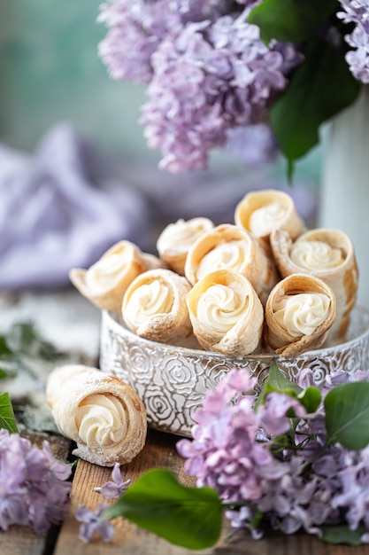 Puff pastry horns with vanilla cream in a metal box in spring still life with a bouquet of lilacs on a wooden table