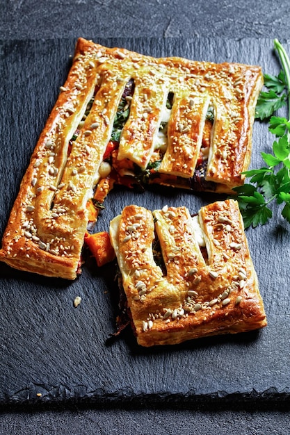 Puff pastry vegetable pie with goat cheese topped with roasted sunflower and sesame seeds filled with roasted vegetables: tomatoes, onion, and pumpkin, on a black slate, top view, close-up