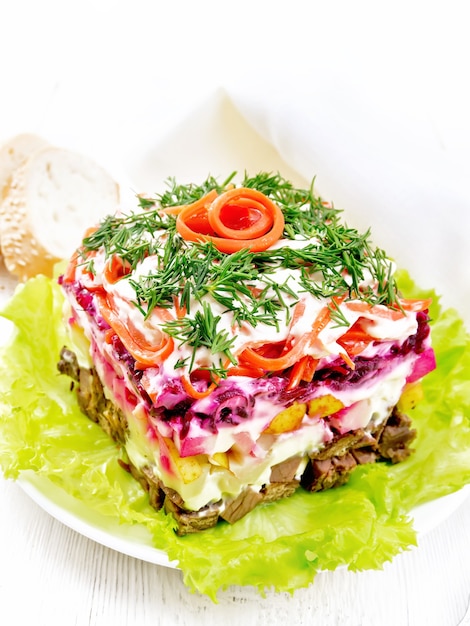 Puff salad with beef, potatoes and beets, pears, Korean spicy carrots, seasoned with mayonnaise and garnished with dill on a green lettuce in a plate, napkin, bread on wooden board background