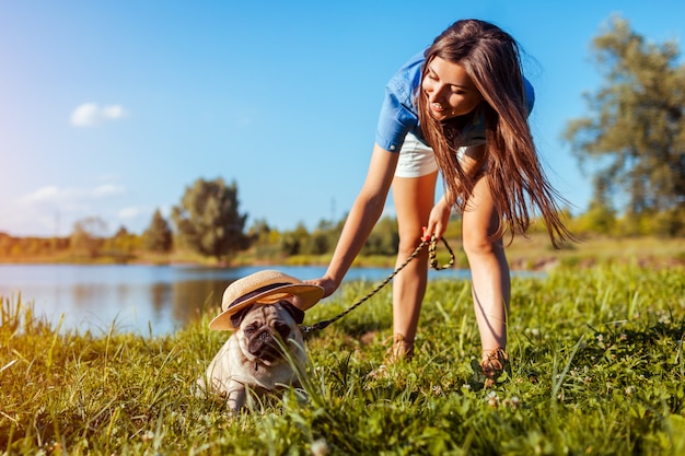 Cane del carlino che si siede dal fiume mentre la donna mette il cappello su. cucciolo felice e suo padrone che camminano e si rilassano all'aperto