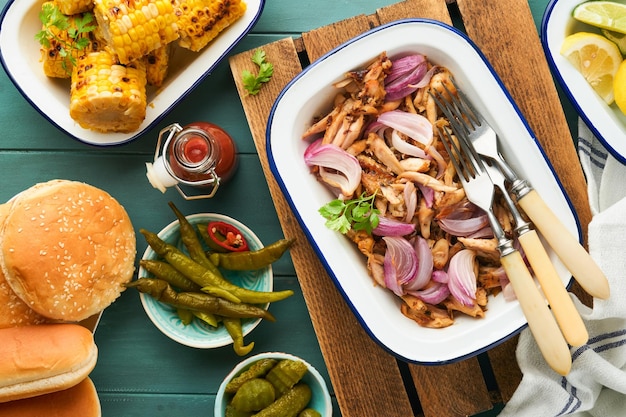 Pulled bbq chicken with baked onions on serving platter bbq corn pickles chili peppers and buns for hot dogs and burgers tomato sauce Traditional American grilled summer bbq sandwiches dishes