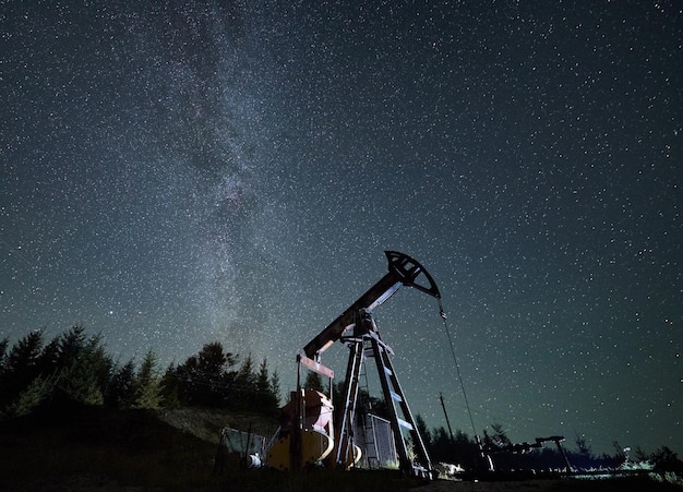 Pumpjack against starry sky with Milky way in natural mountains environment for mining gasoline pipeline system extract crude oil petroleum and water emulsion at each stroke during night time