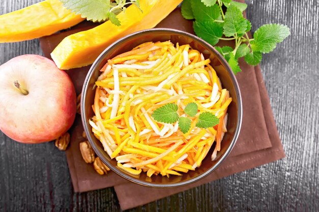 Pumpkin, carrot and apple salad with pecans seasoned with vegetable oil in a bowl on a napkin, mint on dark wooden board background from above