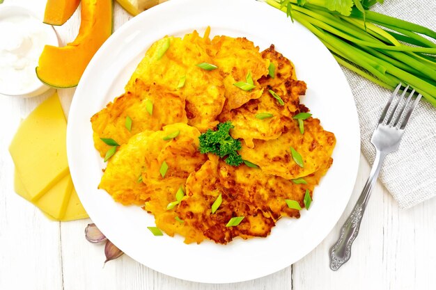 Pumpkin and cheese fritters with ginger in a plate sour cream in a bowl napkin parsley and fork on background of light wooden board from above