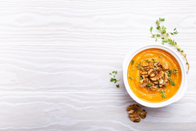 Pumpkin cream soup puree with walnuts, olive oil and herbs in white ceramic bowl on white wooden rustic background, top view, close-up. Delicious vegan soup, space for text