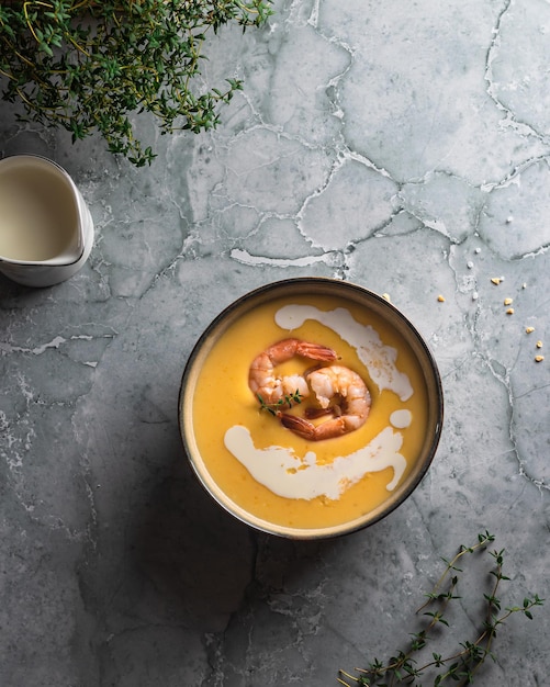 Pumpkin cream soup with shrimp on a marble table thyme is next to the plate The soup is decorated with cream