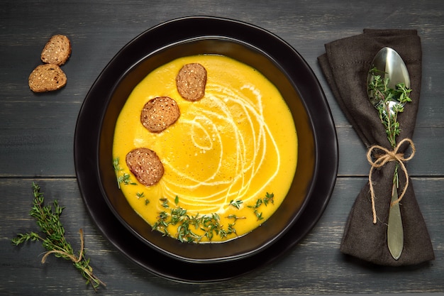 Pumpkin creme soup in ceramic bowl served with croutons, thyme leaves and cream on dark wood