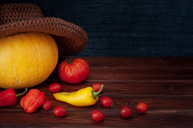 Pumpkin in a hat, scattered red tomatoes and peppers, one yellow pepper. Tomatoes are small and one big. Boards are brown, a background from a scarf fabric. Suitable for halloween. Vignette.