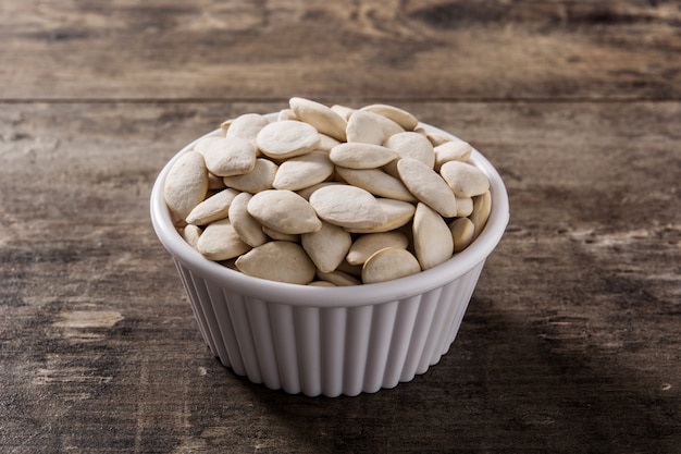 Pumpkin seeds in white bowl on wooden surface.