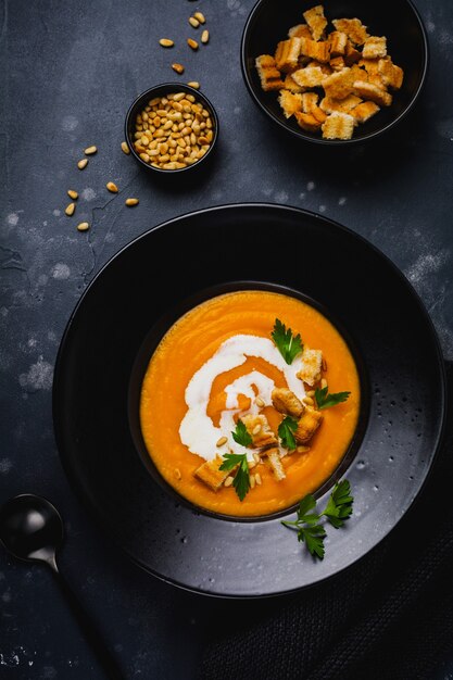 Pumpkin soup with cream, pieces of bread and cedar nuts in black ceramic plate on dark wooden surface
