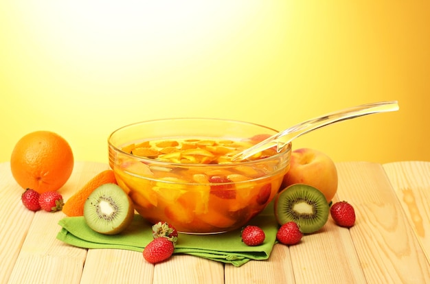 Punch in glass bowl with fruits on wooden table on yellow background