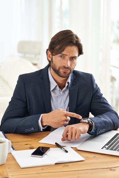 Punctualiteit is cruciaal in dit kantoor. Een knappe zakenman die op zijn horloge wijst terwijl hij aan zijn bureau zit.