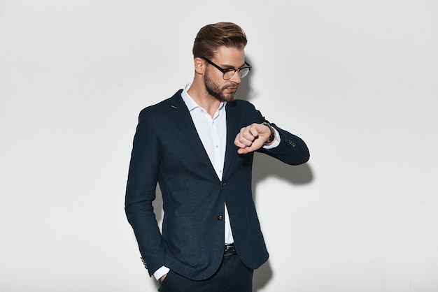 Punctuality is a way to success. Handsome young man in full checking the time while standing against grey background