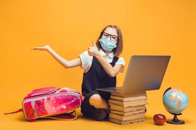 Pupil in medical mask sits behind a stack of books and laptop points the empty space shows thumb up