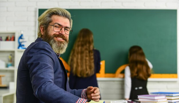 Photo pupil wearing uniform little students studying and reading doing homework writing and reading children girls and teacher man elementary school teacher giving support in classroom selective focus