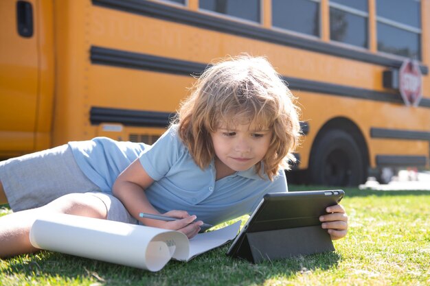 Pupil writing homework on grass with laptop outdoor near school bus distance learning online educati...