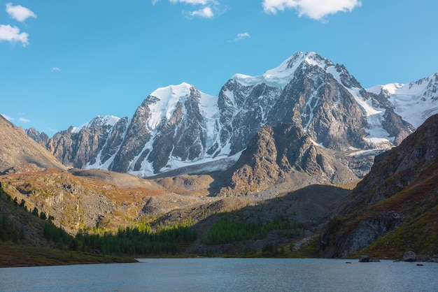 Pure azure alpine lake with view to forest hill and giant snow mountains in autumn sunny day Glacial lake against huge snow covered mountain range in bright sun Vivid autumn colors in high mountains