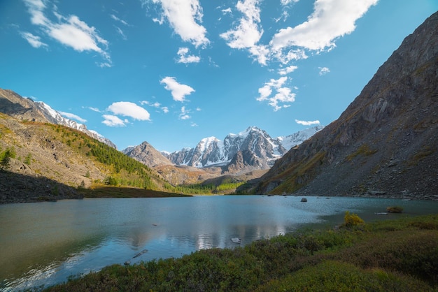 Pure azure alpine lake with view to forest hill and giant snow mountains in autumn sunny day Glacial lake against huge snow covered mountain range in bright sun Vivid autumn colors in high mountains