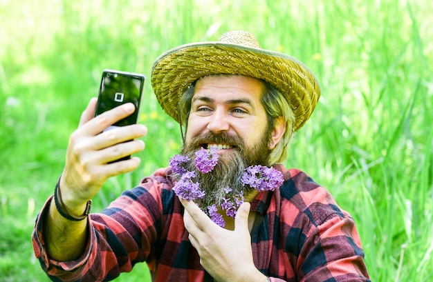 Pure emotions summer selfie with lilac on phone ranch hipster has long healthy beard flower in hair relaxed farmer in straw hat mature man gardener relax on green grass enjoy spring nature