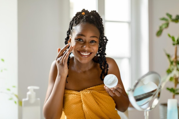 Pure joy of skin hydration smiling black woman applying moisturizing face cream sitting in front of