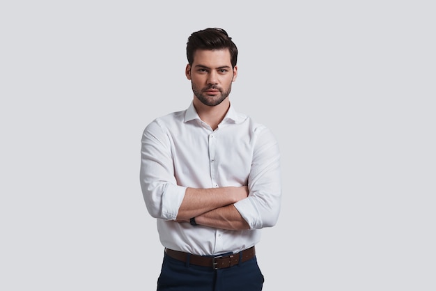 Pure perfection. Good looking young man looking at camera and keeping arms crossed while standing against grey background