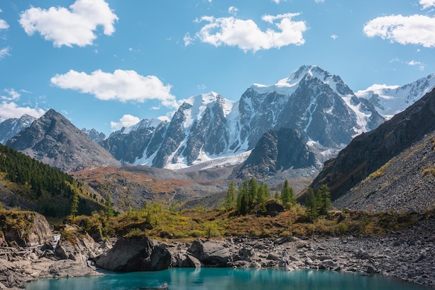 Photo pure turquoise alpine lake with view to forest hill and giant snow mountains in autumn sunny day glacial lake against huge snow covered mountain range in bright sun vivid colors in high mountains