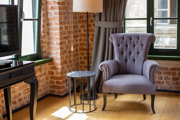 Purple armchair stands near window and tea table in room with red brick walls