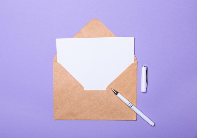 На фиолетовом фоне белая ручка, ремесленный конверт и белая пустая карточка с местом для вставки текста Вид сверху Шаблон