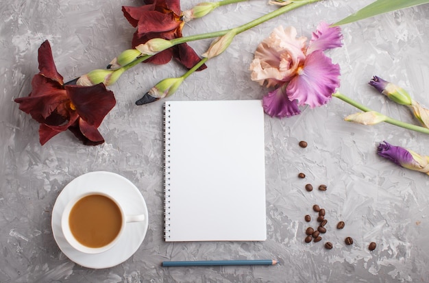 Purple and burgundy iris flowers and a cup of coffee and notebook on a gray concrete background. Morninig, spring, fashion composition. Flat lay, top view, copy space.