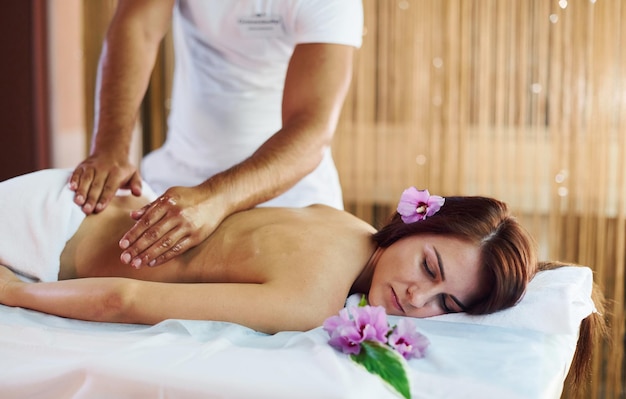 Лежащий фиолетовый цветок Мужчина делает массаж молодой женщине в белом полотенце в помещении