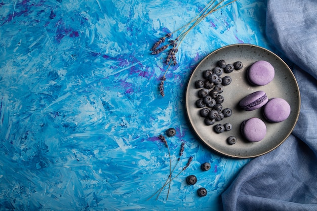 Purple macarons or macaroons cakes with blueberries on ceramic plate on a blue concrete background. 