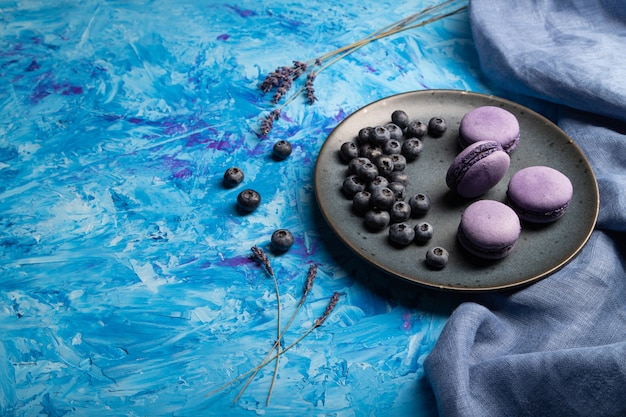 Purple macarons or macaroons cakes with blueberries on ceramic plate on a blue concrete background. 